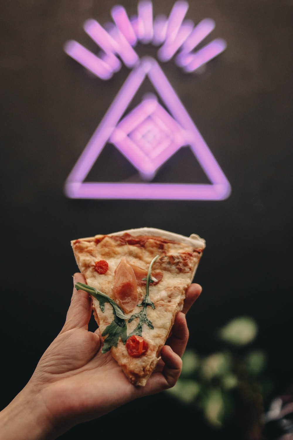 a hand holding a slice of pizza in front of a neon sign