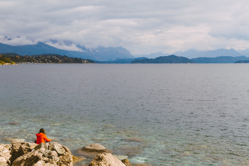 person in orange jacket sitting on rock near body of water during daytime
